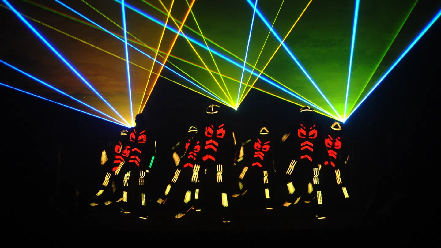 Tron dancers in red and yellow LED suits on a dark stage under fans of blue, green and yellow laser beams
