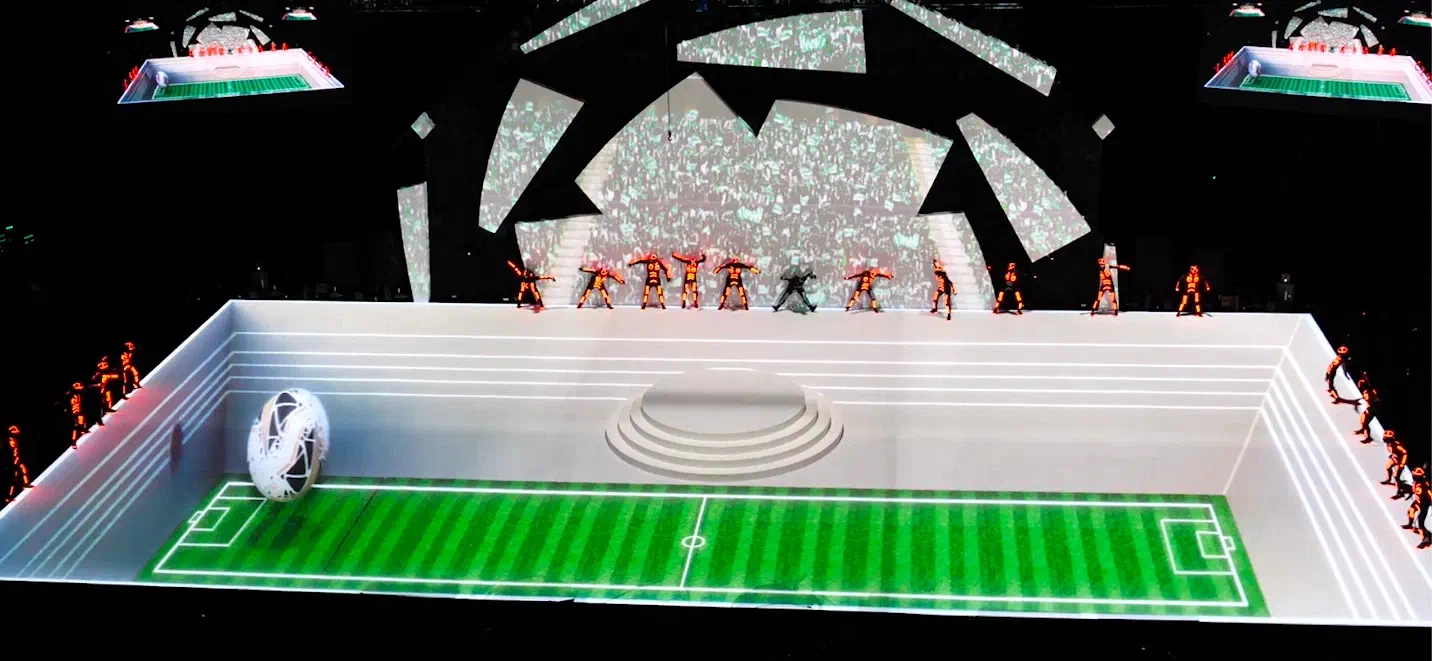 Tron dancers in orange LED suits standing on top of a white box-shaped stage with a football pitch and a giant ball projected onto it, images of a cheering crowd projected behind them and two LED screens above
