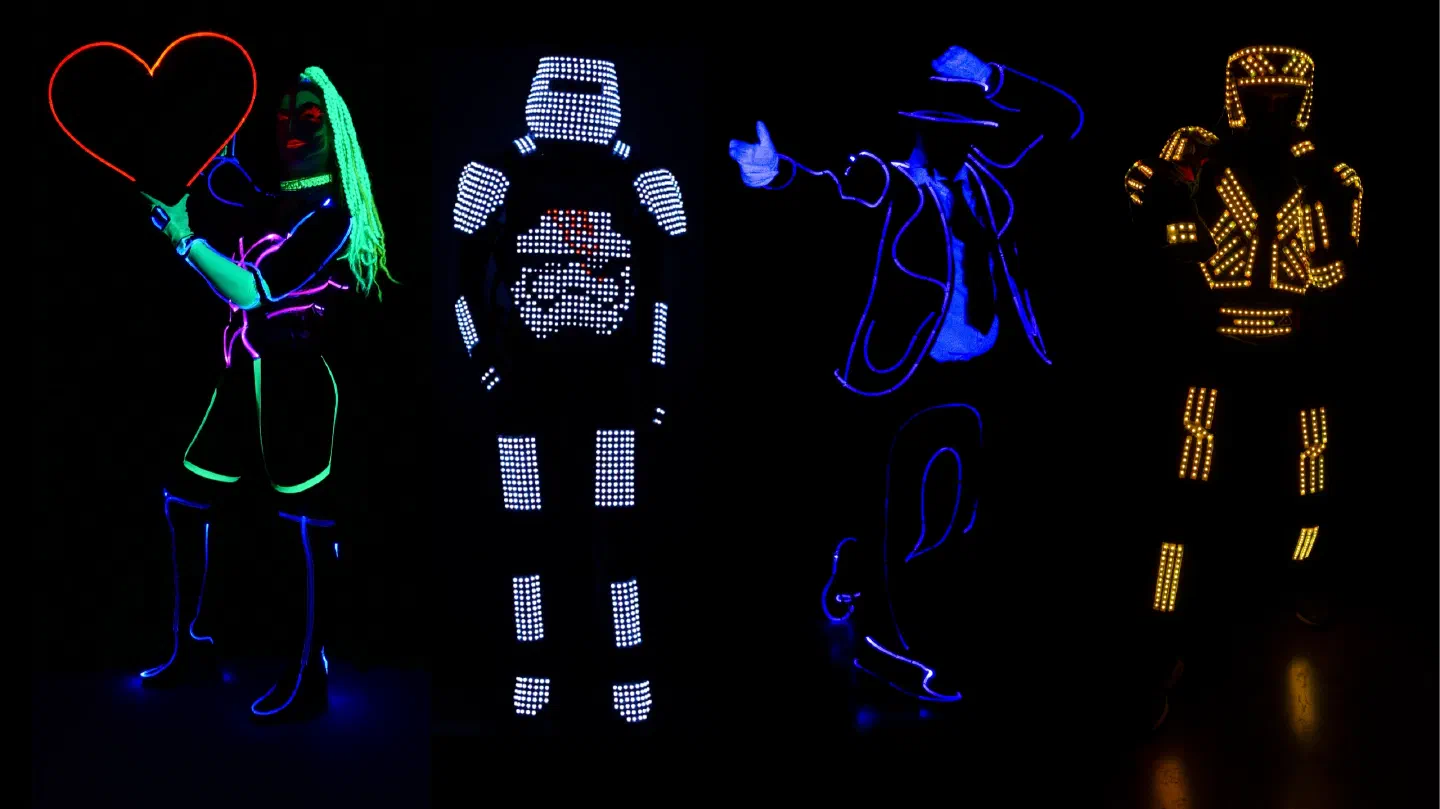 Four of our LED suits glowing in the dark – a dancer with UV face paint and glowing green hair holding a red neon heart, a pixel suit of white LEDs with a stormtrooper helmet on its chest, a dancer in a hat outlined in blue light and a suit with rows of yellow LEDs