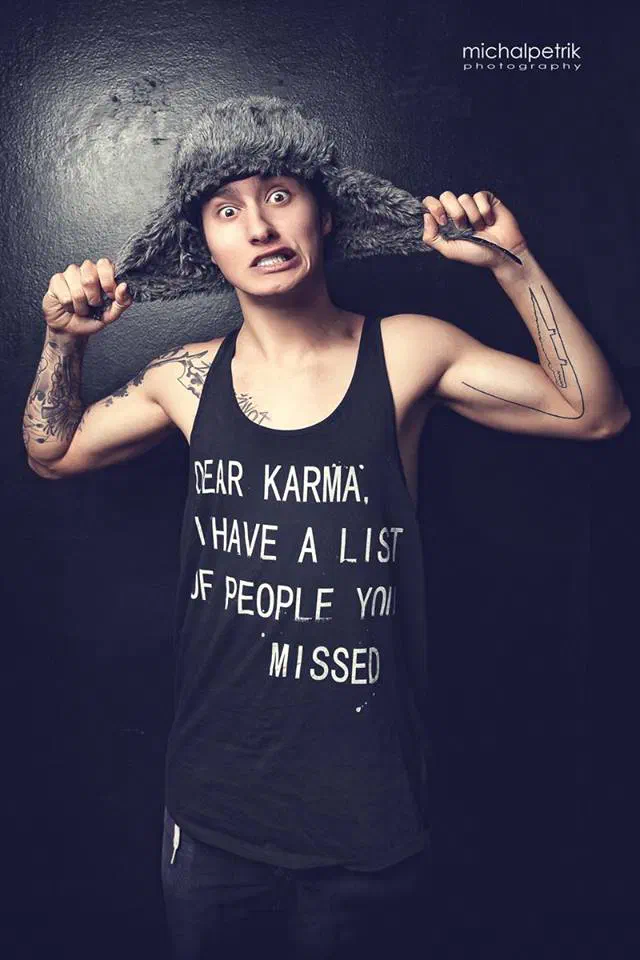 Tony, our dancer – portrait of a young man in a black tank top that reads “Dear Karma, I have a list of people you missed”, holding out the ear flaps of a fur hat and pulling a face