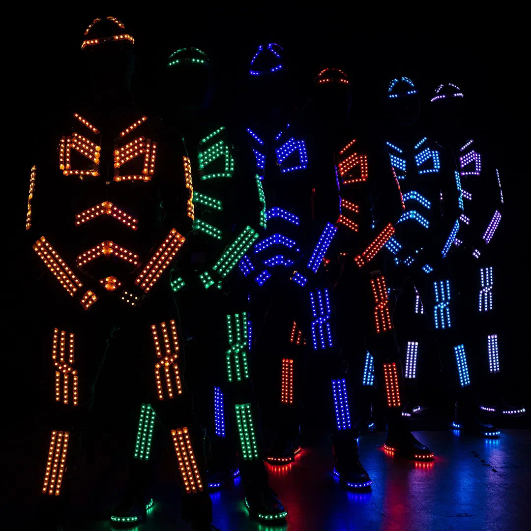 Šesť tanečníkov v LED kostýmoch Tron Dance stojí v rade v tme, segmenty LED pásikov na prilbách, hrudi, rukách a nohách žiaria oranžovo, zeleno, modro, červeno, svetlomodro a fialovo