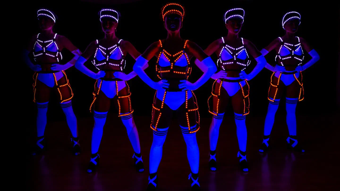 Päť tanečníc Tron Dolls v bielych kostýmoch žiariacich pod UV svetlom namodro, s bielymi a oranžovými LED postrojmi, čiapkami a klietkovými sukňami, stoja v tme s rukami vbok