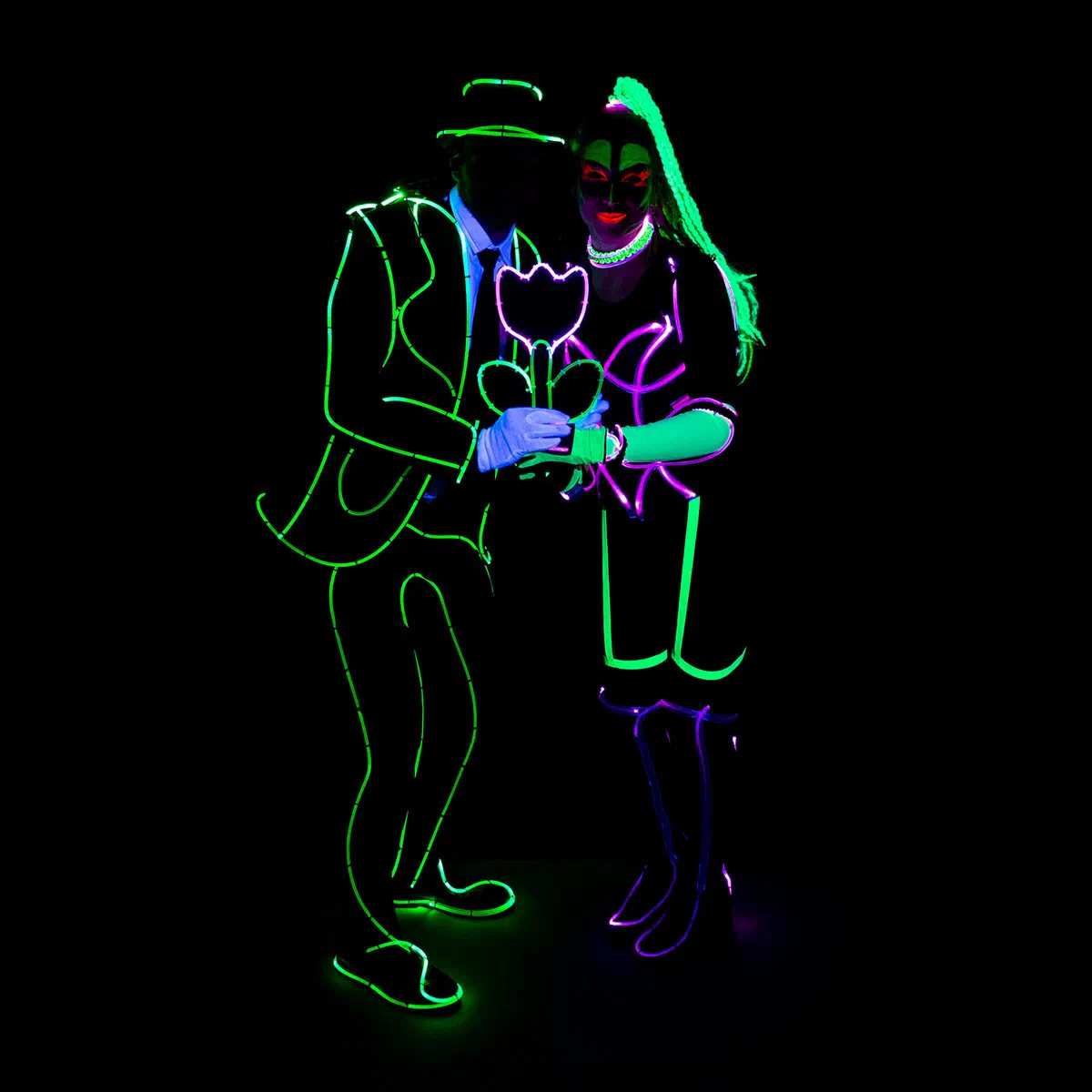A male dancer in a glowing green outline suit and hat and a female dancer in a glowing UV costume hold a pink and green LED flower together