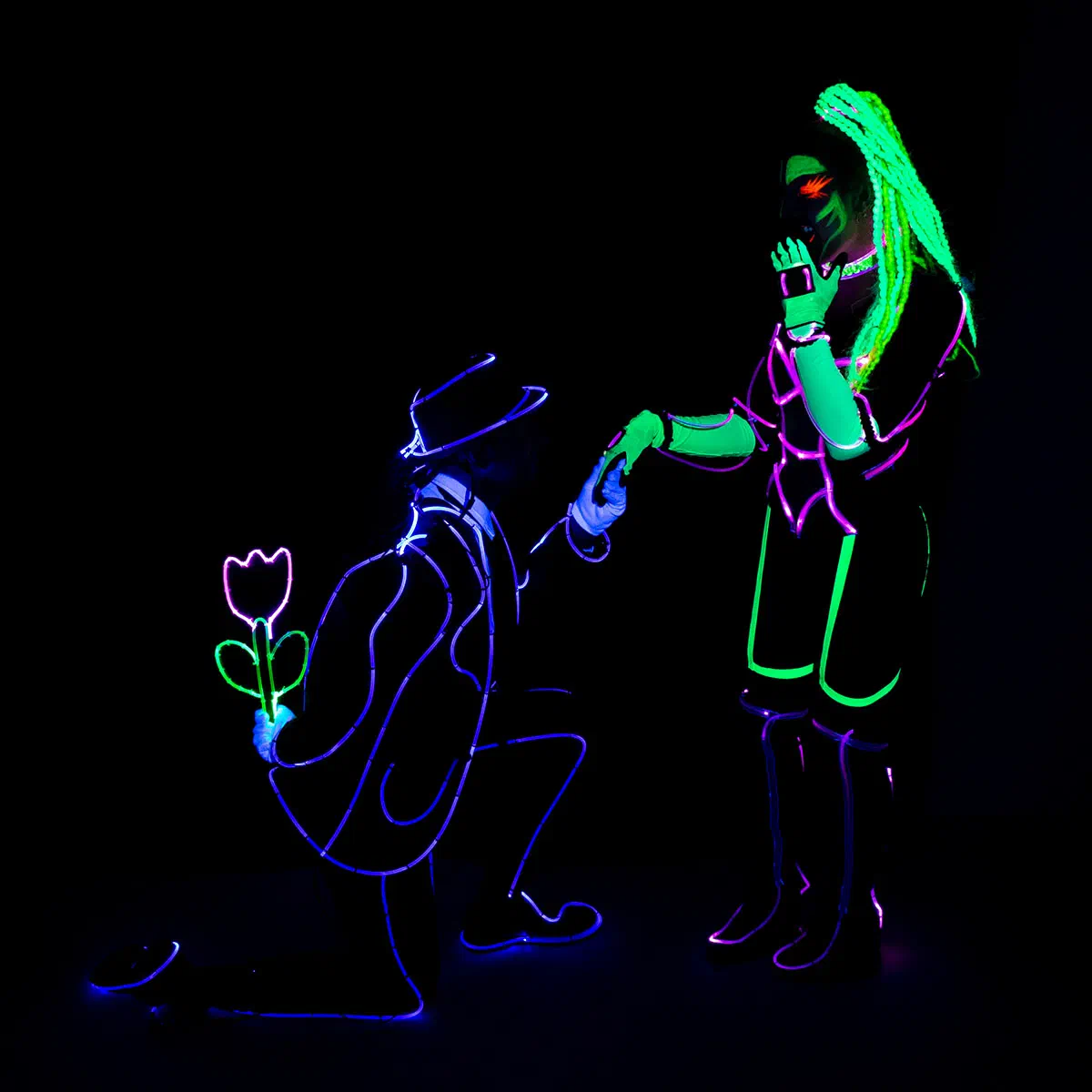A male dancer in a glowing blue outline suit and hat kneels with an LED flower behind his back and holds the hand of a female dancer in a green and purple glowing UV costume with neon braids
