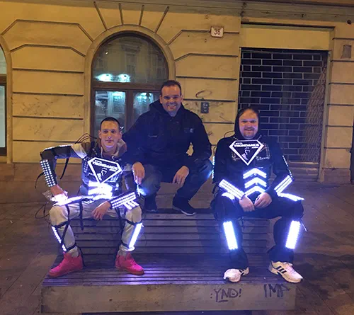 Two of our technicians in LED suits with the glowing Tron Dance logo sign on the chest, sitting on a street bench at night with a third man in a dark jacket between them