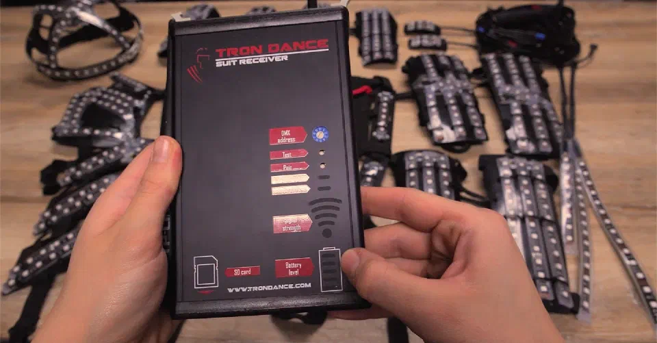 Tron Dance suit receiver held in two hands – a black box with DMX address, test, pair, signal strength, SD card and battery level markings – above the LED strip parts of a suit on a wooden table