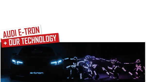 Audi E-Tron + our technology – red and white banners above a dark Audi e-tron with glowing blue headlights, next to a row of Tron dancers in glowing blue and purple LED suits bent forward as if pushing the car, in the dark
