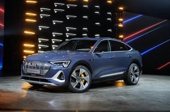 Blue Audi E-Tron electric SUV with a sloping roof on a dark presentation stage, in front of a wall of glowing white light bars and red and blue light lines