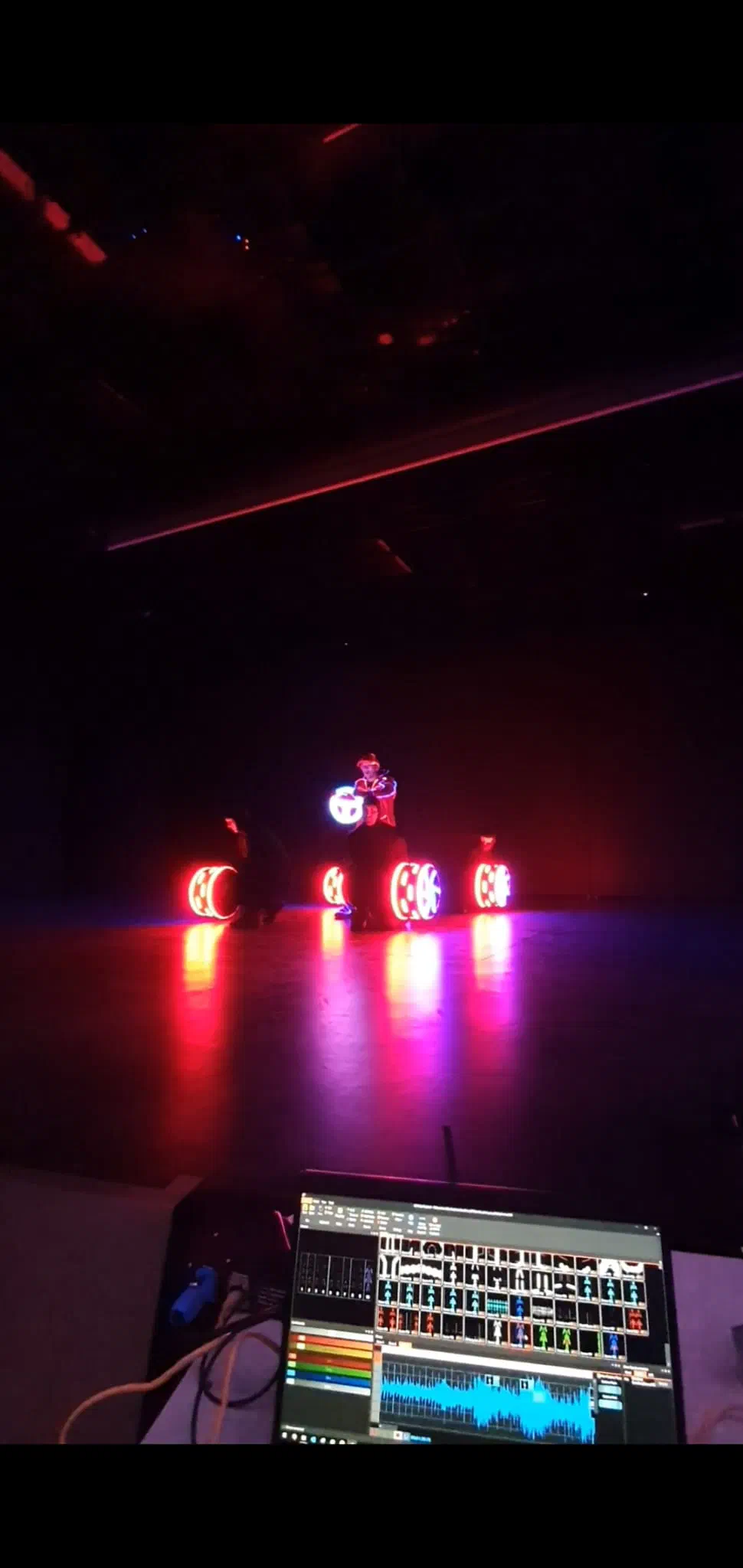 Four LED wheels glowing red on a dark stage around a performer holding a lit LED steering wheel, a laptop with the show programming in the foreground