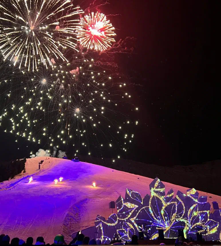 Fireworks burst over the Serfaus ski slope at night – the snow lit purple, small fires along the slope, and the stage’s crystal-shaped panels glowing with projected golden patterns above the audience