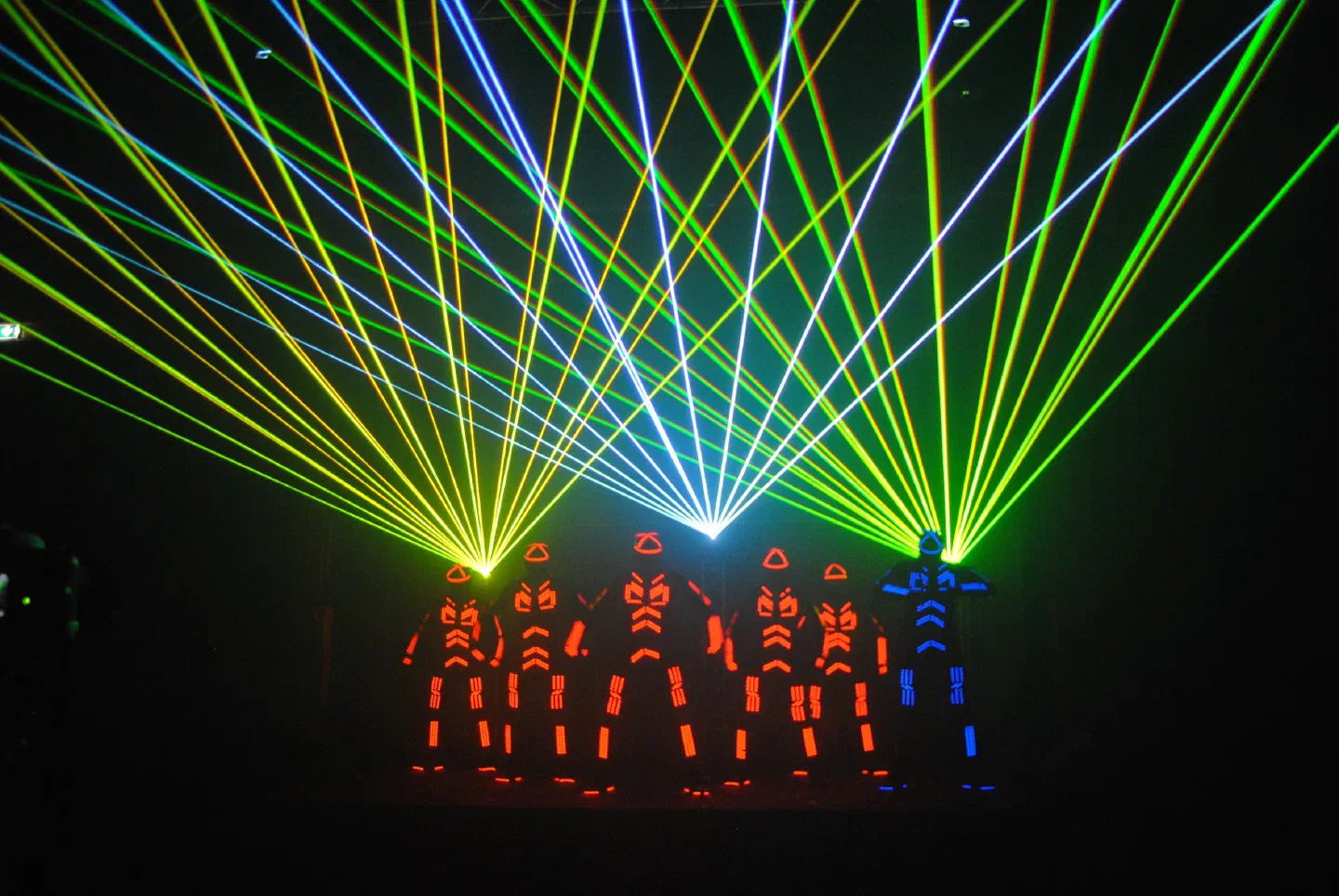 Six Tron dancers in orange and blue LED suits on a dark stage under a fan of green, yellow and white laser beams