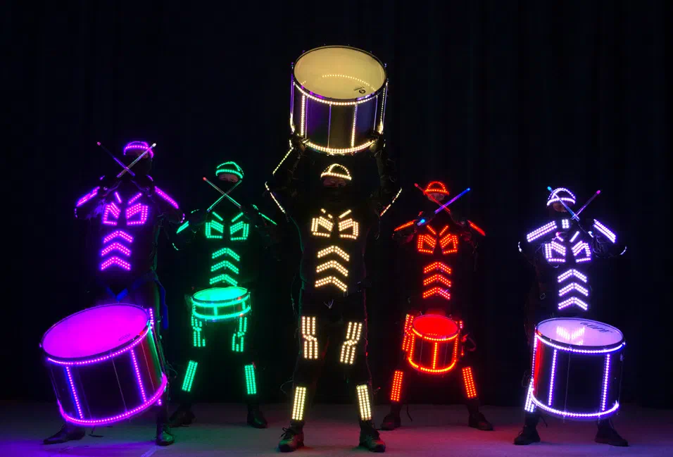 Five Tron Drummers in glowing violet, green, yellow, red and white LED suits with illuminated drums, the one in the middle holding his drum overhead