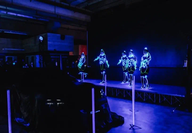 Five Tron Girls in glowing LED suits dancing on a blue-lit stage, a covered car and light poles in the foreground