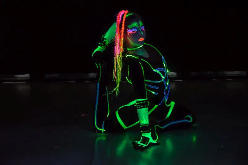 A Tron Girl kneeling on the stage floor in a suit with curved green and blue fibre optic lines, with neon UV make-up and pink braids