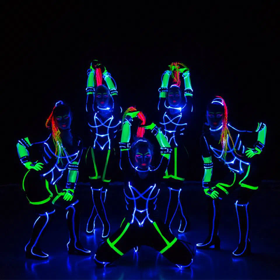 Five Tron Girls posing on a dark stage in black suits outlined with glowing blue-white fibre optic cable, with neon green UV sleeves and red braided hair