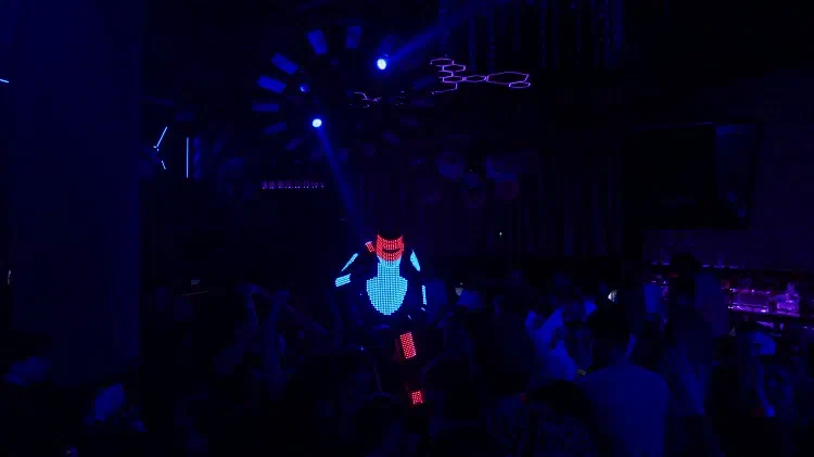 Tron Pixel Robot stilt walker with a red pixel helmet and a glowing blue pixel chest towering over a dancing crowd in a dark club lit by blue beams