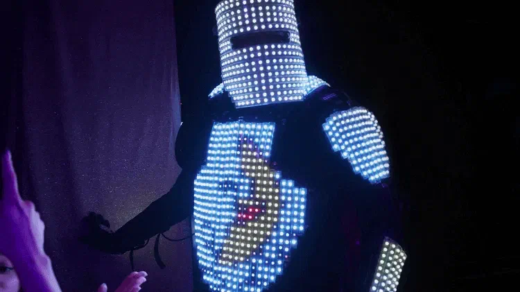Tron Pixel Robot close up – a helmet and chest made of white and blue LED pixels, the chest showing a colourful pixel picture, a guest’s hand reaching towards it