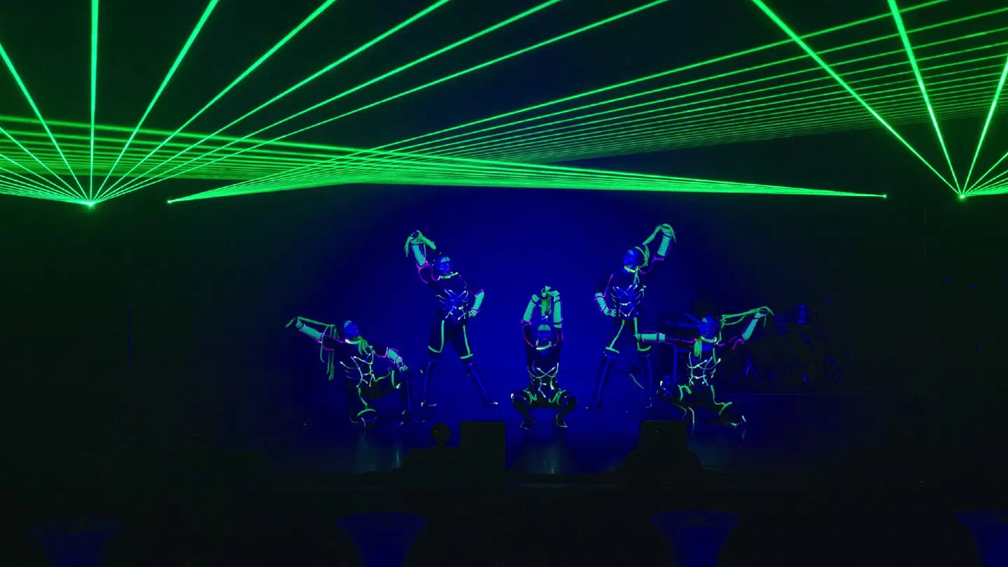 Five Tron Girls in glowing LED costumes posing on a blue-lit stage under fans of green laser beams