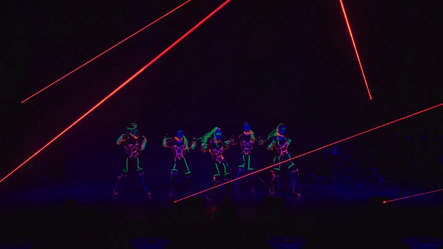 Five Tron Girls in glowing pink and green LED costumes dancing on a dark stage, crossed by red laser beams