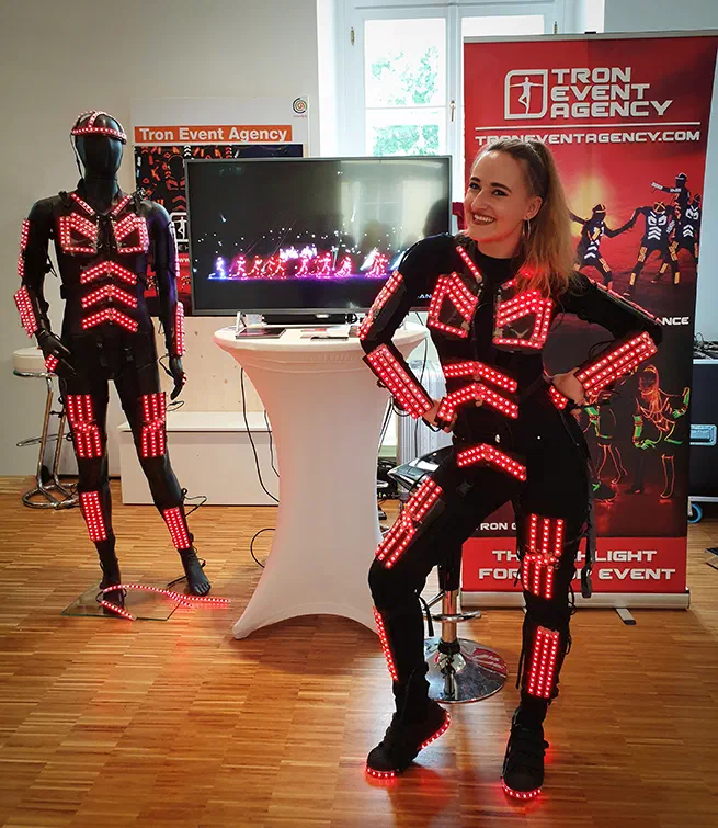 A smiling dancer in a black LED suit with glowing red LED chevrons at our stand, next to a mannequin in the same LED suit, a screen showing an LED dance show and a Tron Event Agency roll-up banner