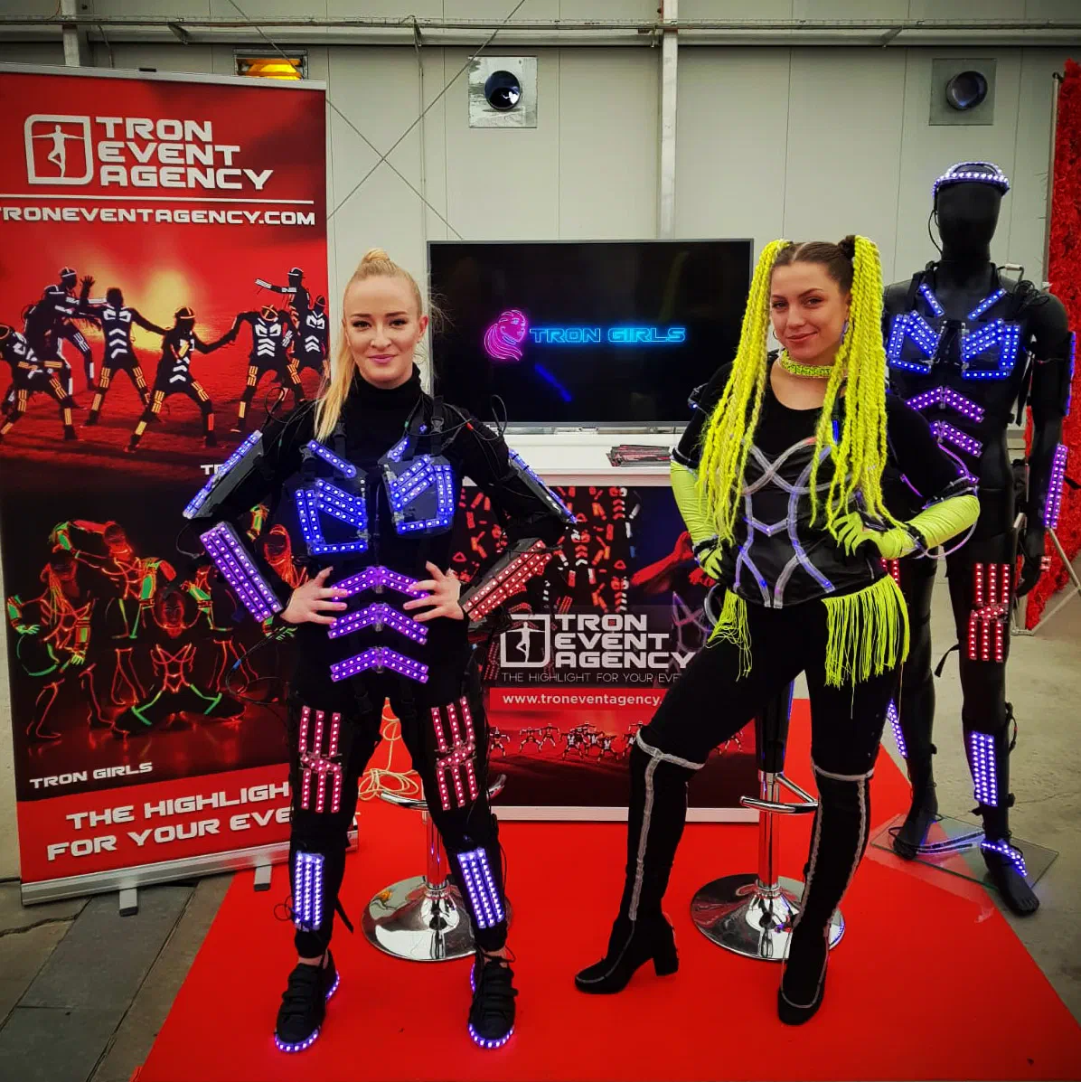 Two dancers – one in an LED suit with glowing blue and pink stripes, one in a black costume with neon yellow braids – at our Tron Event Agency stand, with a Tron Girls screen, roll-up banners and an LED suit on a mannequin