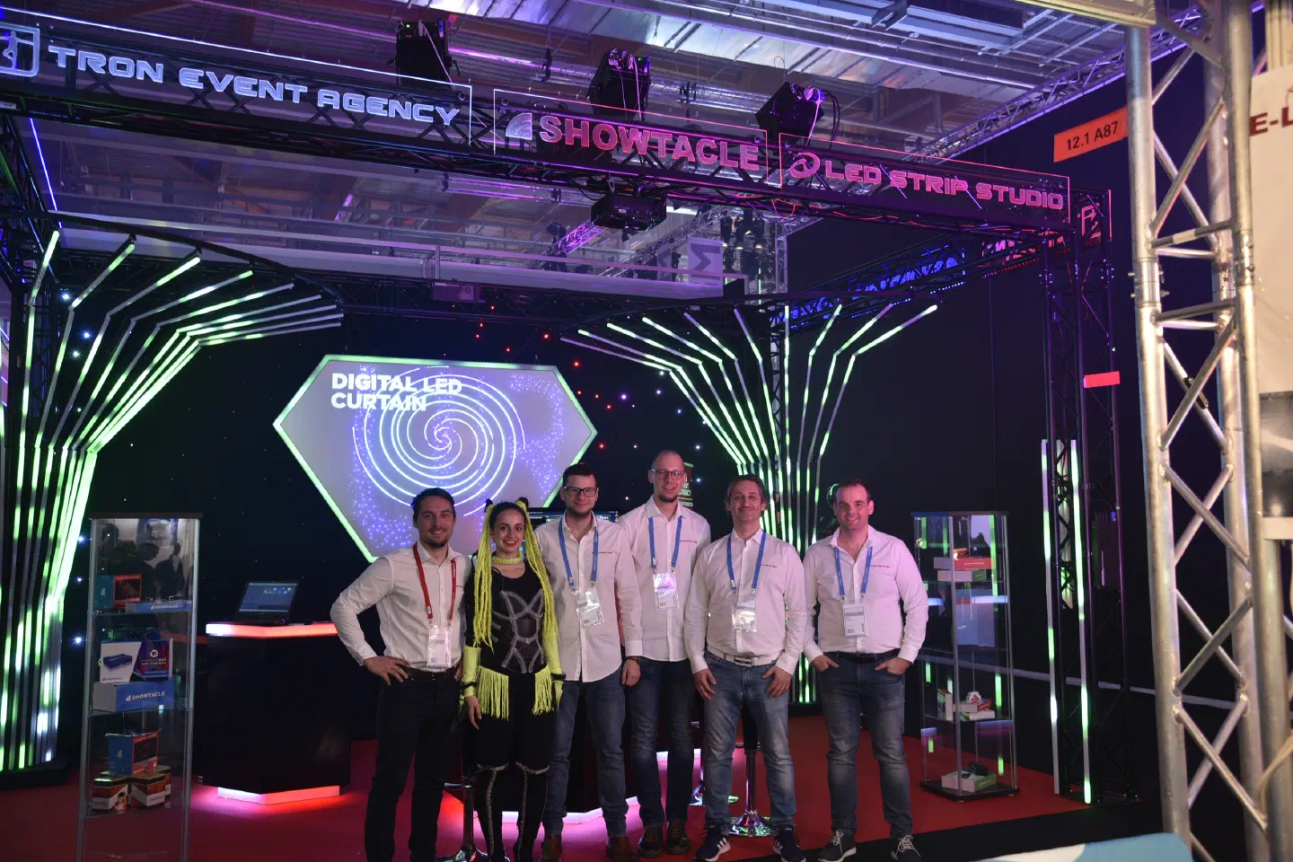 Our team – five men in white shirts and a dancer in a black costume with neon yellow braids – at our Pro Light & Sound stand under Tron Event Agency, Showtacle and LED Strip Studio signs, with glowing green LED tubes and a Digital LED Curtain screen