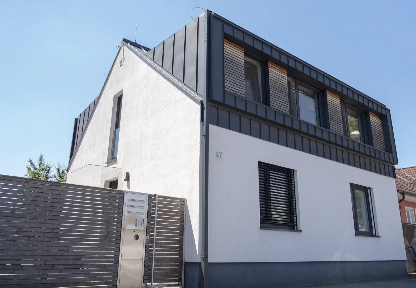 Our office building in Bratislava on a sunny day – a white house with a dark metal-clad upper floor and wooden window slats, behind a wooden fence with a steel mailbox gate