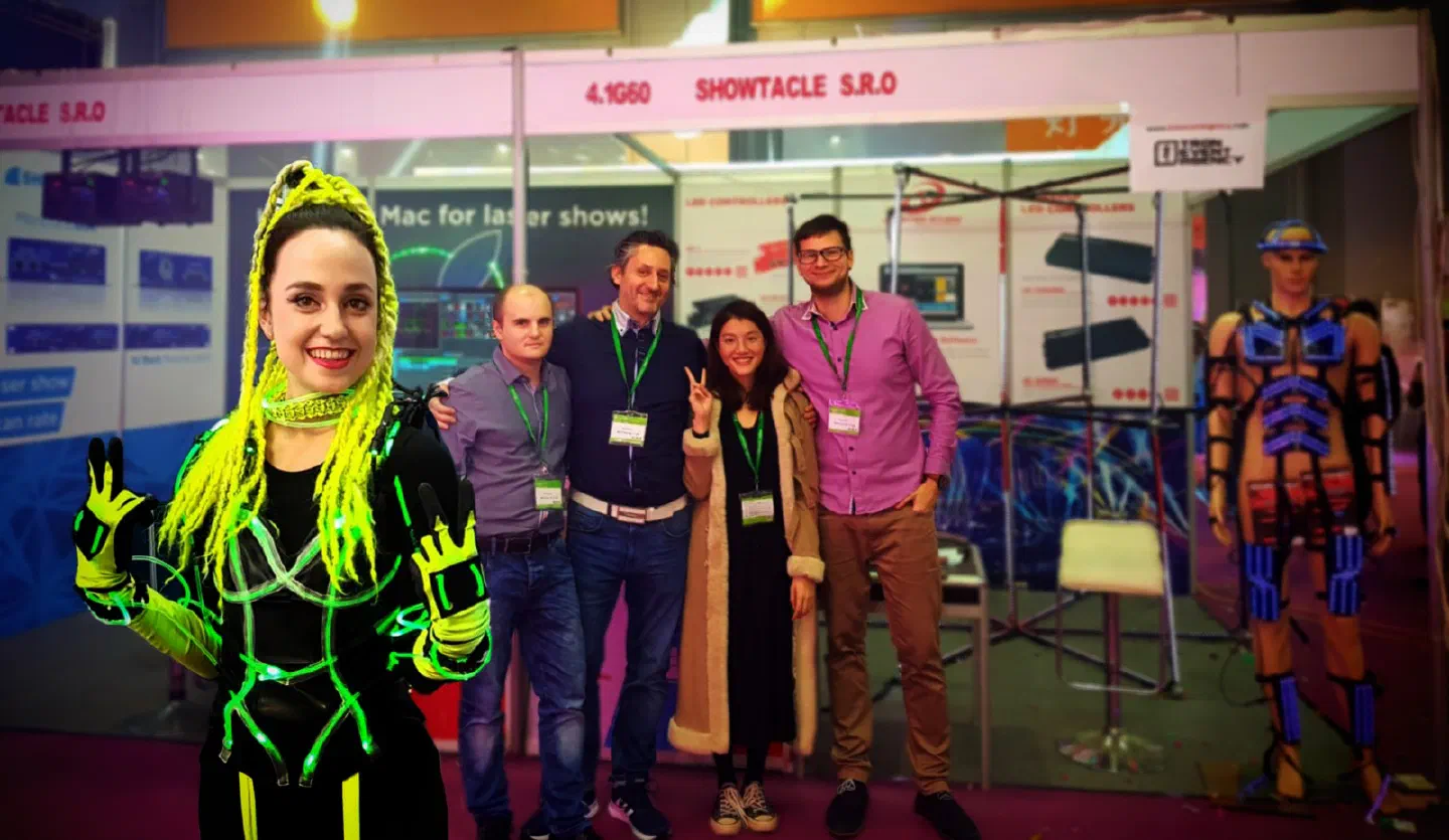 Our team and a dancer in a glowing green LED costume with neon yellow braids at the Showtacle s.r.o. booth 4.1G60, with LED controller posters behind them and a mannequin in a blue LED suit on the right