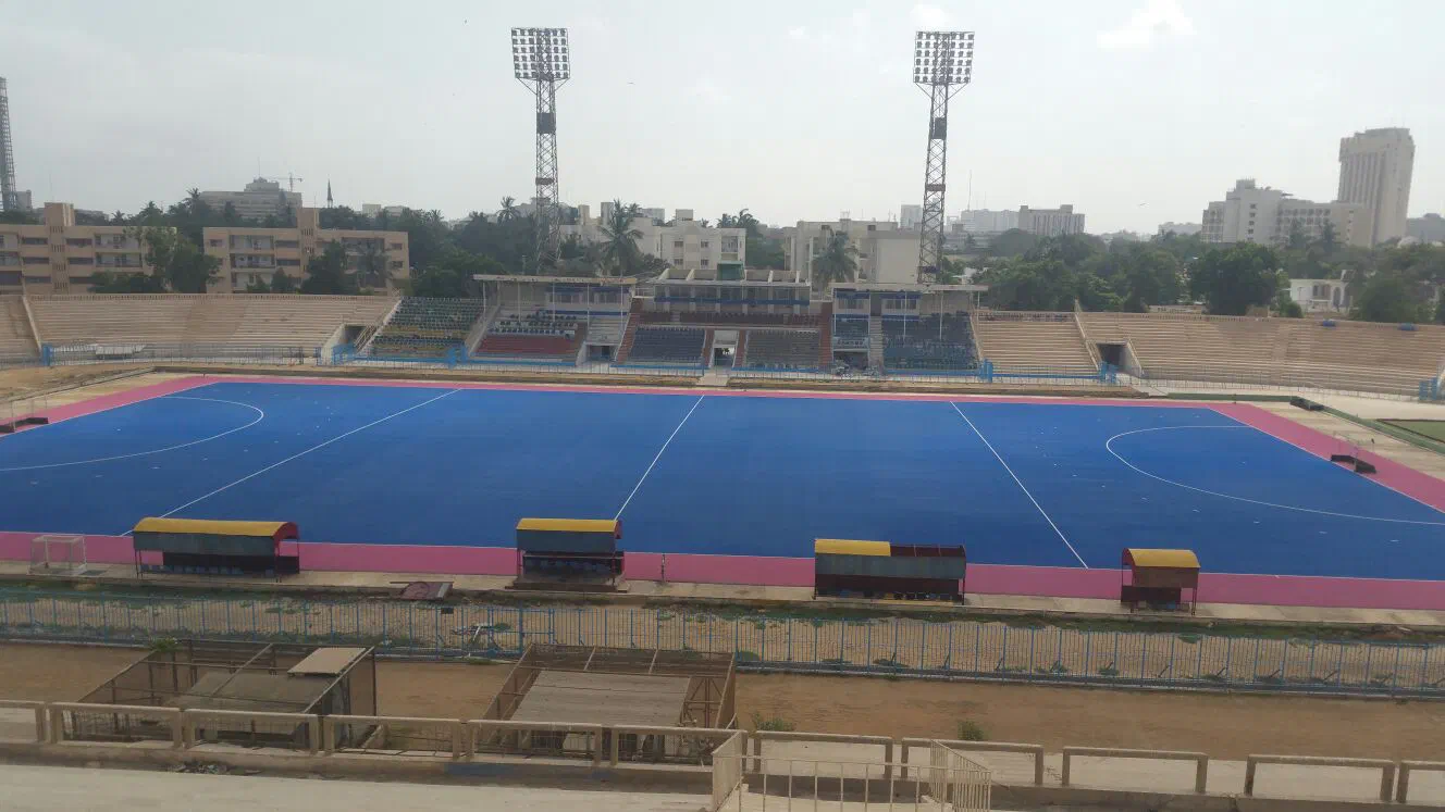 Empty hockey stadium in Karachi by day – a blue pitch with pink edges, floodlight towers and stands, with city buildings and palm trees behind it