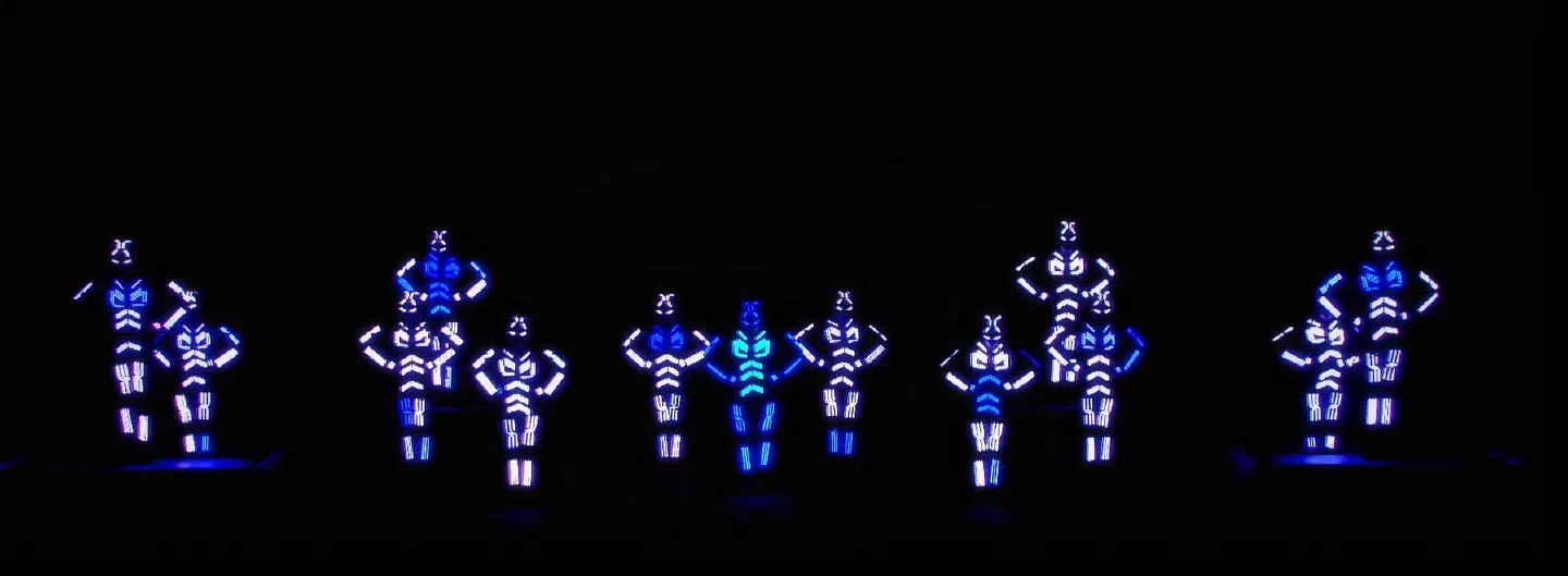 A big LED dance crew spread across a dark stage in small groups, their LED suits glowing white and blue, the one in the middle turquoise