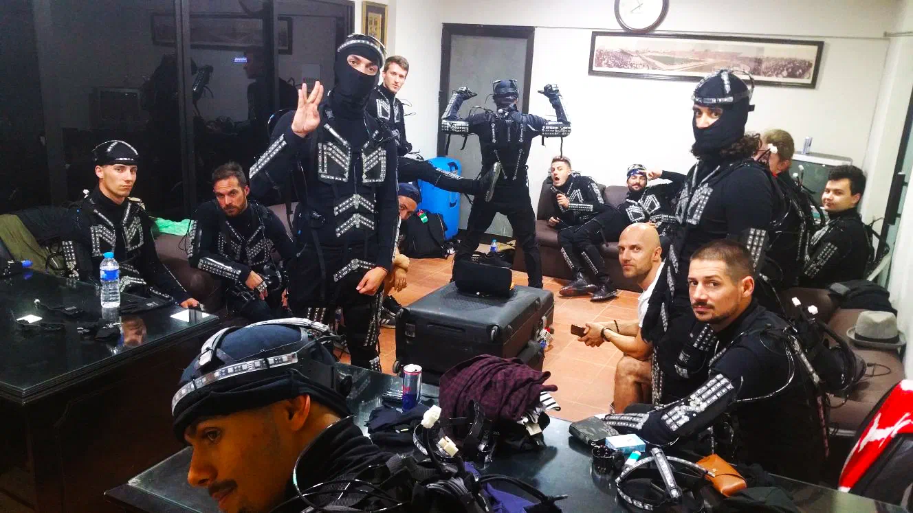 Our team backstage in a dressing room – dancers in black LED suits, some with LED helmets and masks, around a table covered with cables and LED helmet parts, a hard-shell suitcase beside it