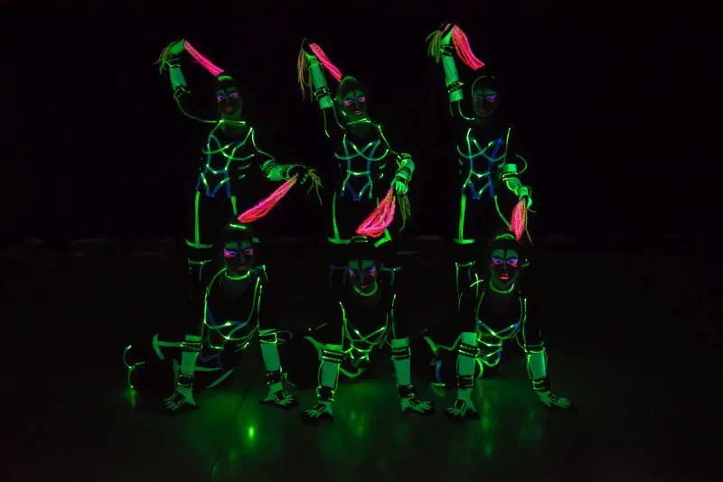 Six female dancers posing in two rows on a dark stage, their costumes lined with glowing green and blue stripes, with UV face paint and pink tassels in their hands