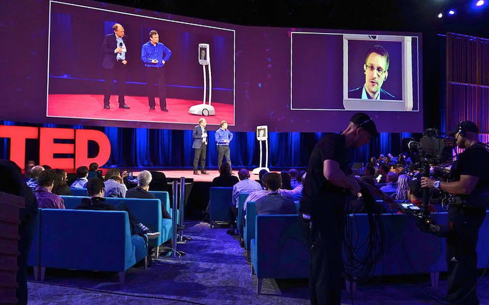 A TED conference stage with the big red TED letters: two speakers stand next to a telepresence robot while large screens show them and the face of a man taking part remotely, with the audience in blue armchairs and two cameramen filming in the foreground