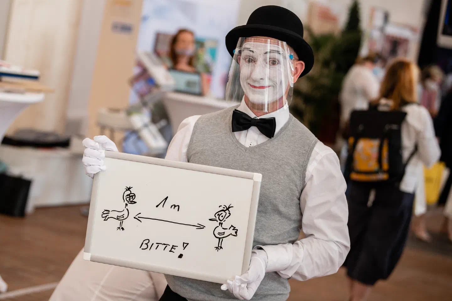 A mime in white face paint, a bowler hat and a face shield holds a small whiteboard asking for 1 m distance – two drawn birds with an arrow between them and the word Bitte!