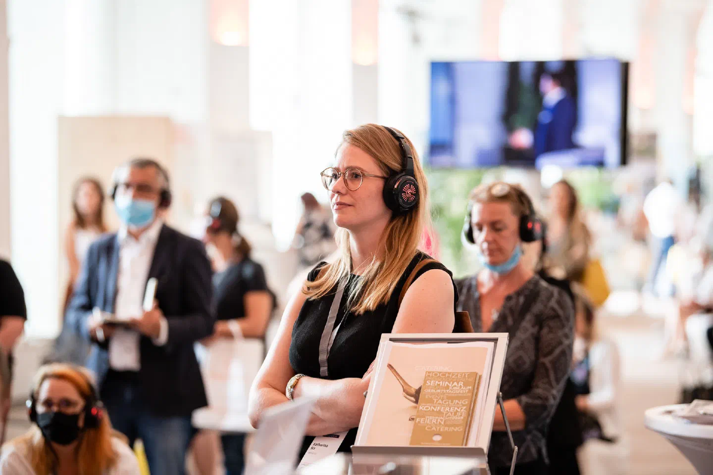 Visitors with wireless headphones, some of them in face masks, listen to a seminar in the Eventbiz hall, with a screen showing the speaker in the background