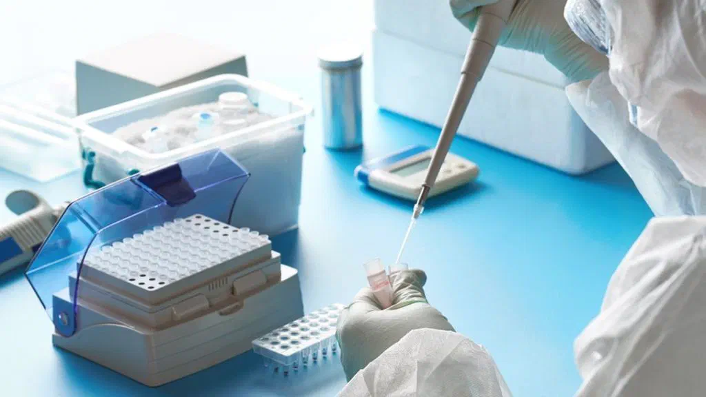 A laboratory worker in a protective suit and gloves pipettes a sample into small test tubes next to a rack of tubes on a blue table