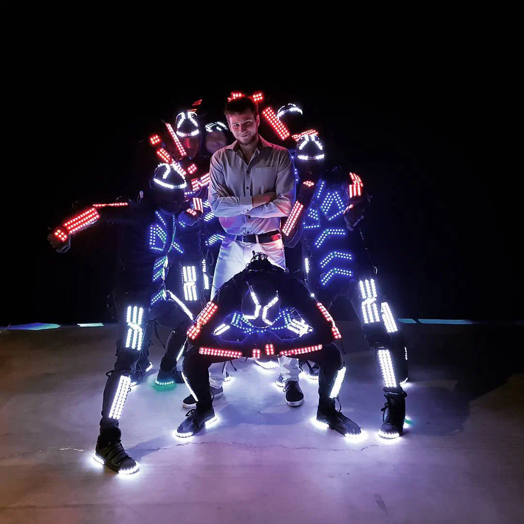 Richie in a light shirt with folded arms surrounded by Tron dancers in black LED suits glowing red, white and blue in the dark