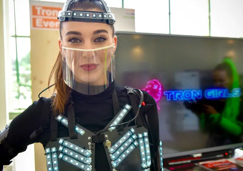 Tron dancer in a black LED suit with white LED stripes, an LED headband and a clear face shield at our stand at the Eventbiz exhibition, in front of a screen with the neon Tron Girls logo