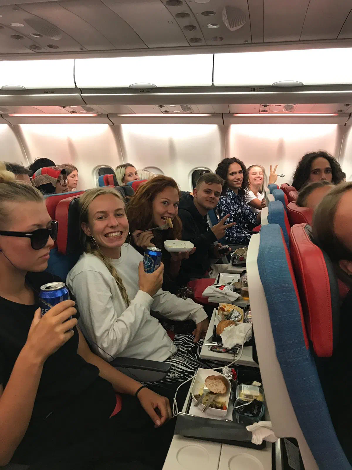 Smiling Tron dancers in their seats on a plane with meal trays and drinks, some of them waving and showing the peace sign to the camera