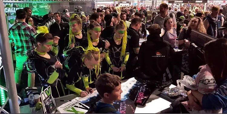 Tron Girls with neon yellow braids and green face paint, in black costumes with yellow sleeves, sign autographs for fans at a crowded stand in Birmingham