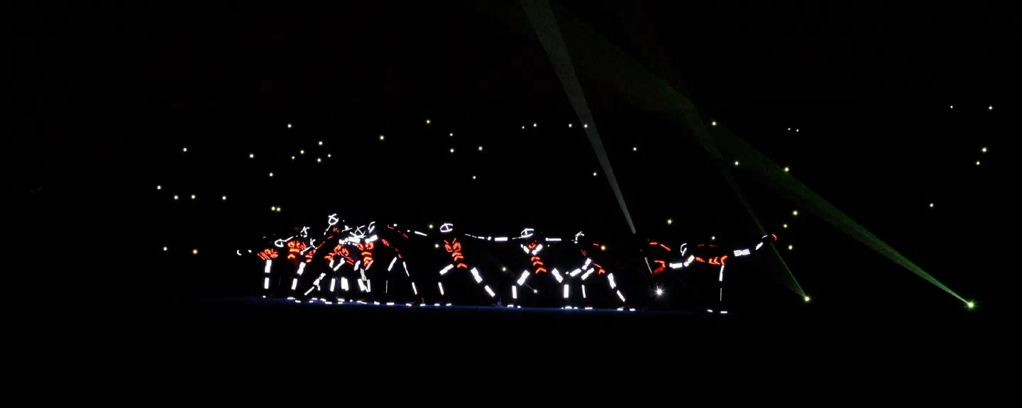 A line of dancers in LED suits glowing white and orange on a dark stage, with green laser beams from the right and phone lights of the audience behind