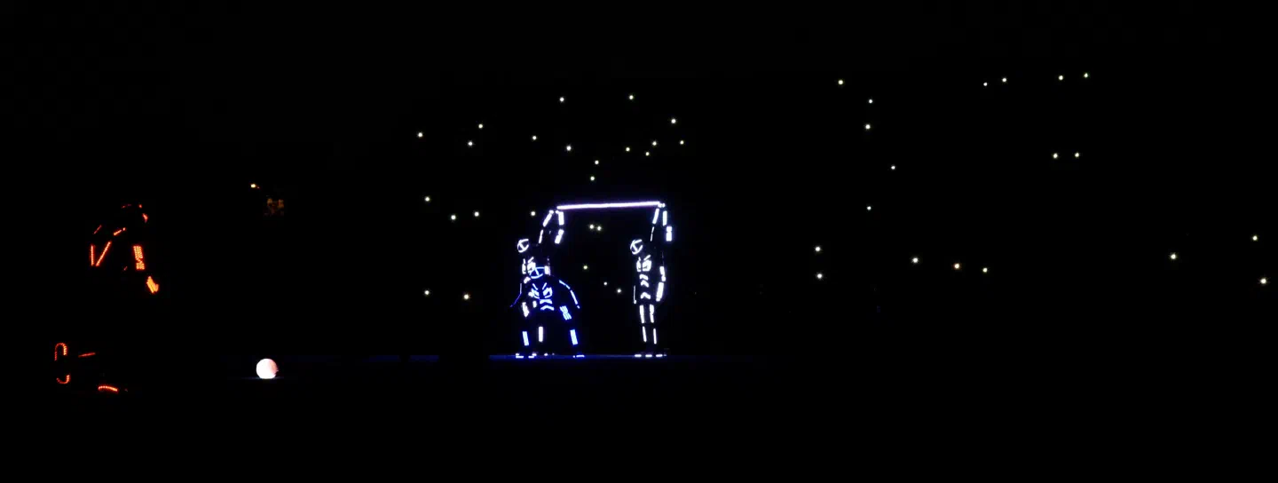 Dancers in LED suits acting out a football scene in the dark – two blue and white figures form a goal with a glowing crossbar while an orange-lit player stands by a glowing ball, with phone lights of the audience behind