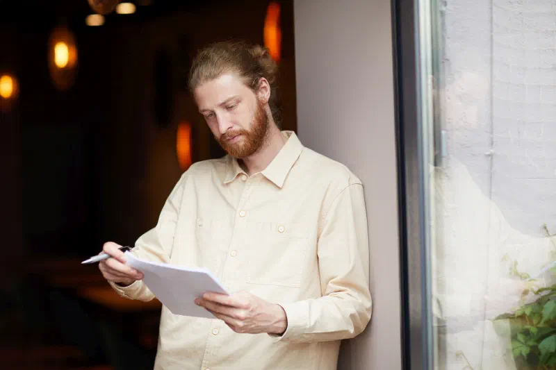 A bearded man in a light shirt leaning by a window and reading notes on a sheet of paper with a pen in his hand