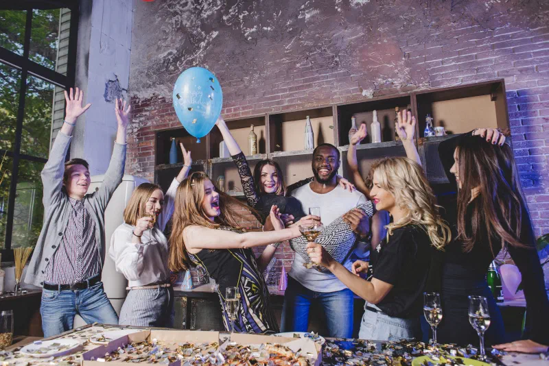 A group of young friends celebrating at a home party – raised hands, a blue balloon, glasses of sparkling wine and a table covered in confetti