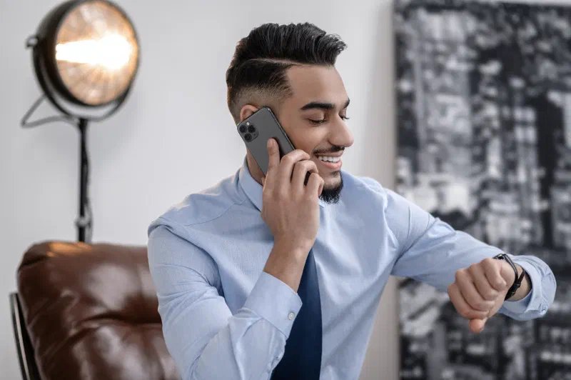 A smiling young man in a light blue shirt and tie talking on the phone while checking his watch