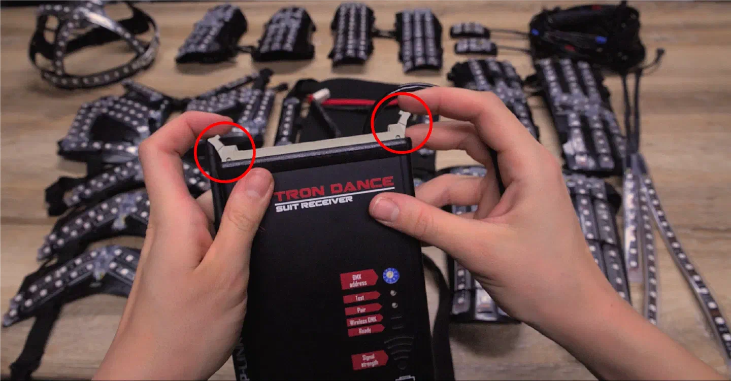 Hands holding a Tron Dance suit receiver with the two plastic lockers on its top edge circled in red, in front of LED suit parts laid out on a wooden table