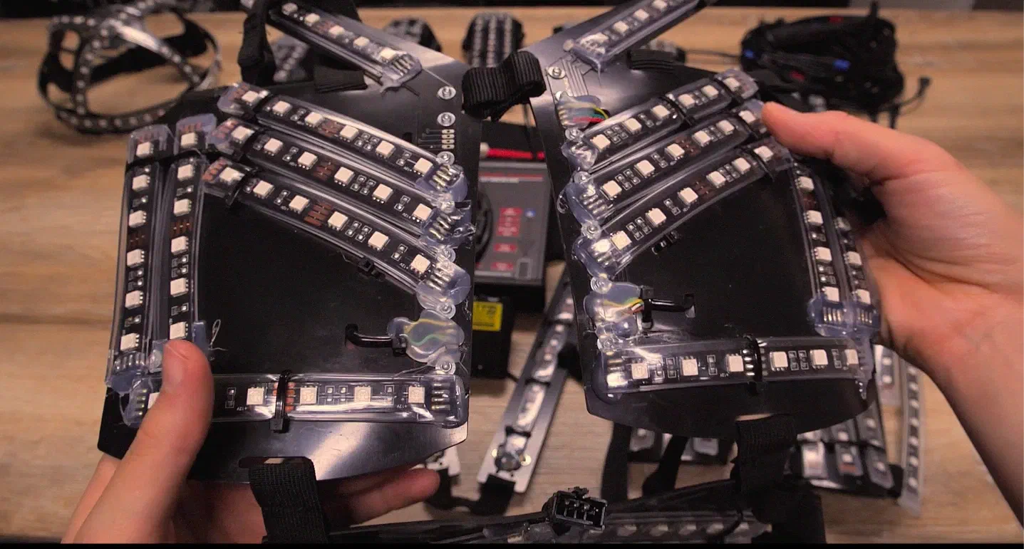 Hands holding two chest parts of a Tron Dance suit – rows of LEDs sealed in clear plastic tubes on black plastic plates – above the other suit parts on a wooden table