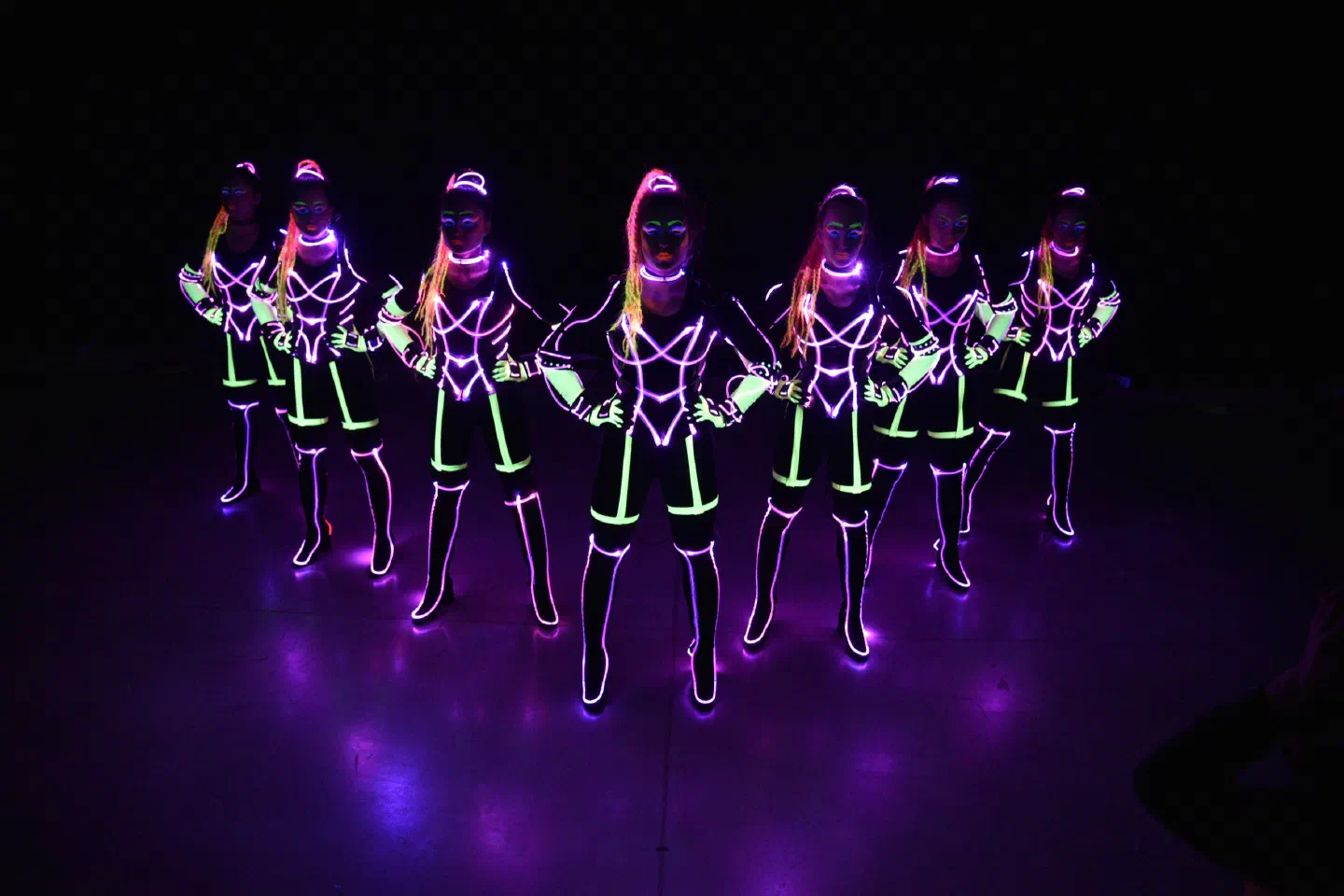 Seven Tron Girls dancers in black costumes with glowing pink and neon green lines, standing in a row on a dark stage