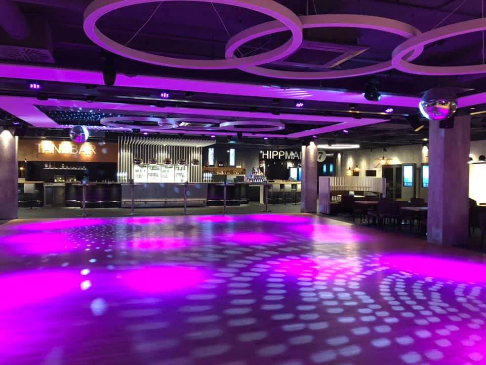 Empty event hall before the show – a large dance floor lit purple and dotted with mirror-ball light, a long bar at the back and white ring lights hanging from the ceiling