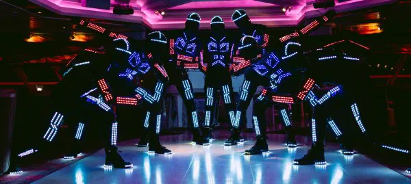 Tron dancers in black suits lit by blue, white and red LED strips, posing together on a glowing dance floor under pink lights